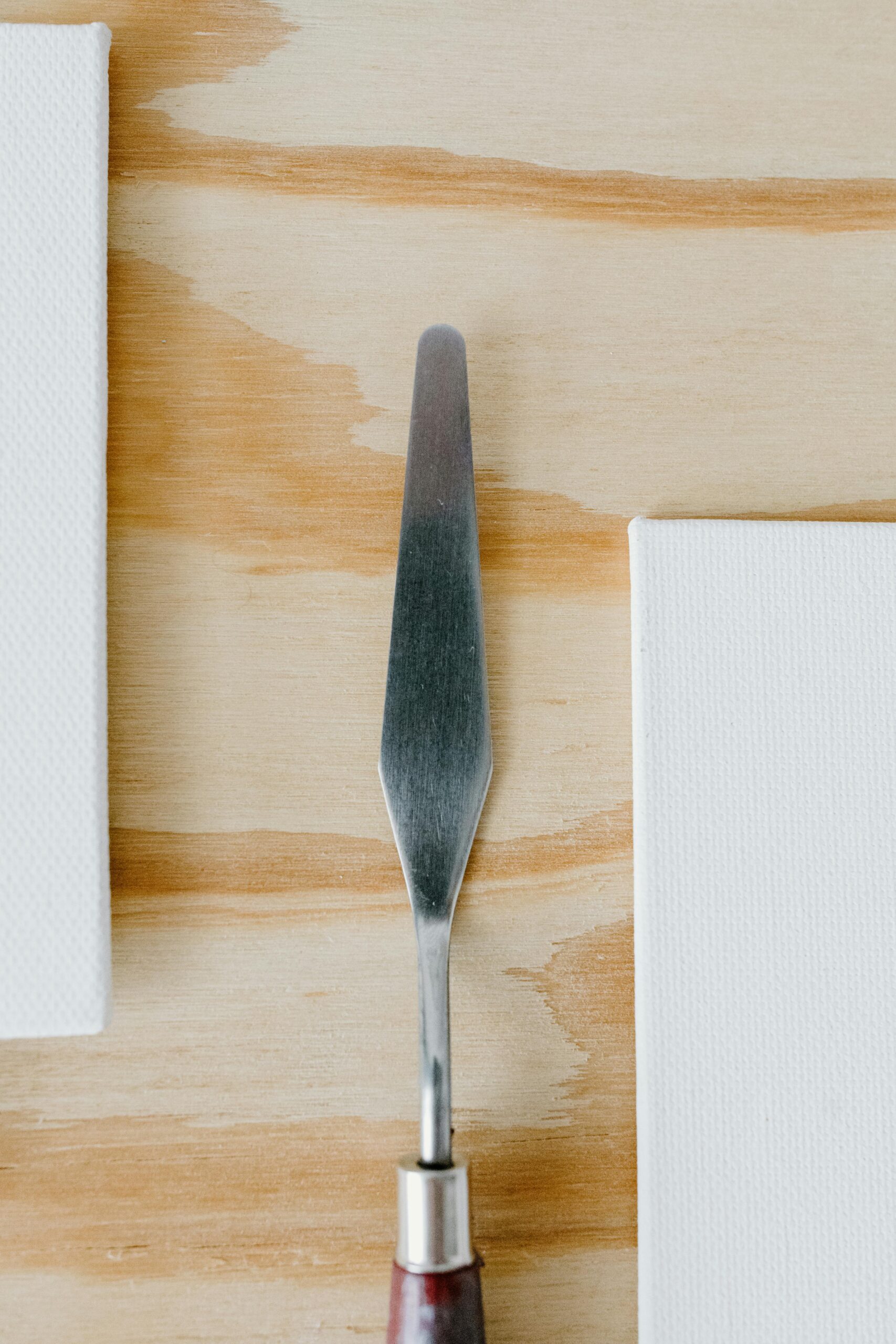 Palette knife placed on a wooden desk between two blank canvases, ready for painting.